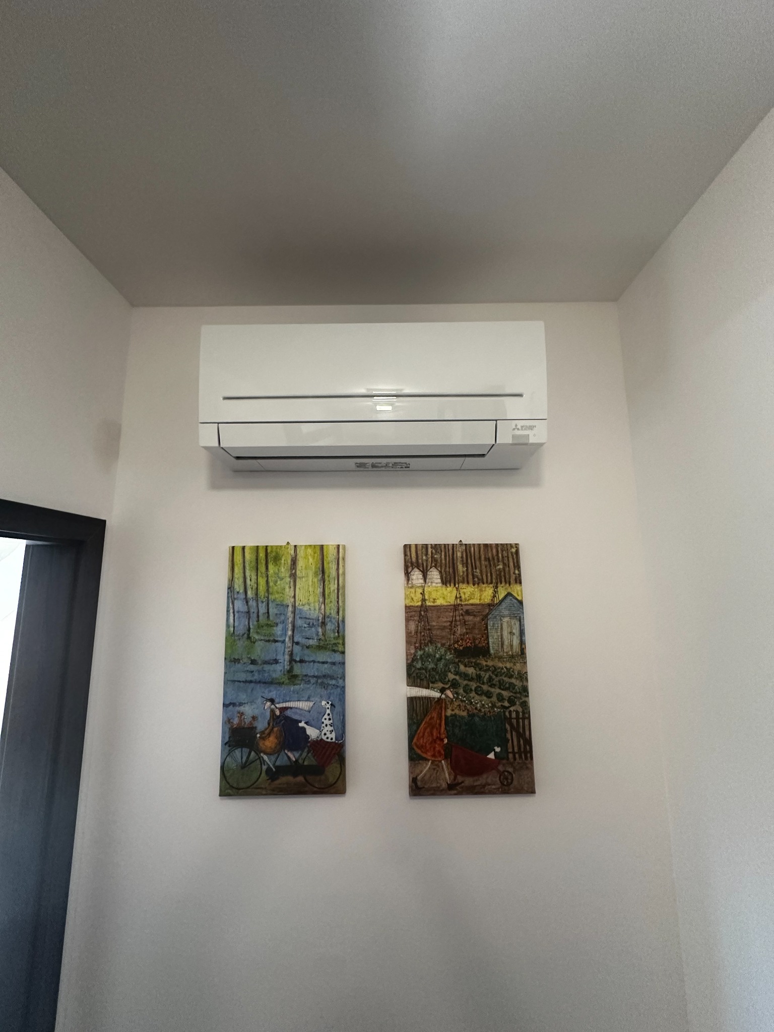 Biały klimatyzator ścienny marki Mitsubishi, zamontowany w rogu pokoju nad dwoma obrazami o tematyce wiejskiej.