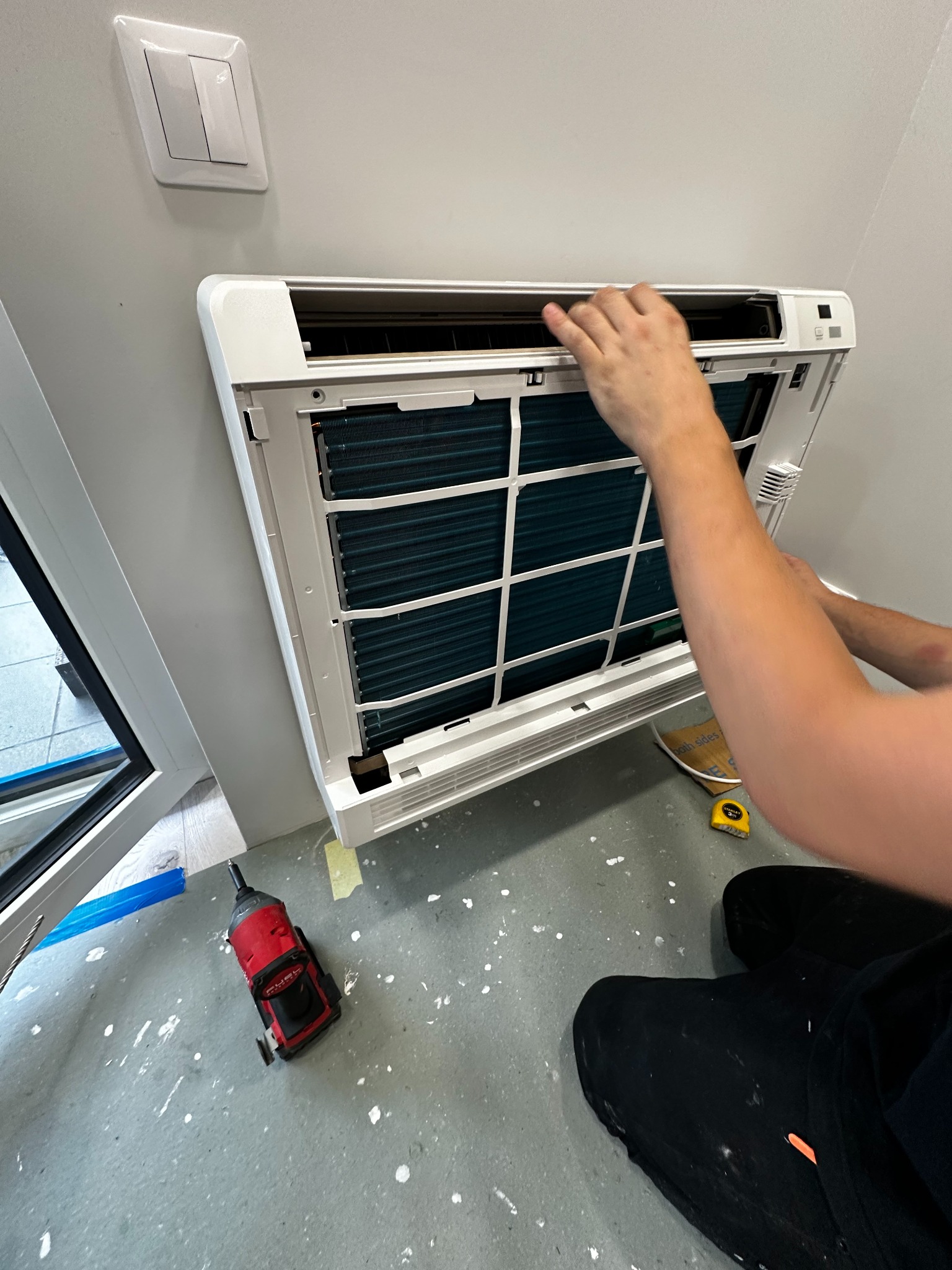 Instalacja białej klimatyzacji ściennej w pomieszczeniu, widoczne ręce montera, wkrętarka udarowa i miarka leżące na podłodze z plamami farby.