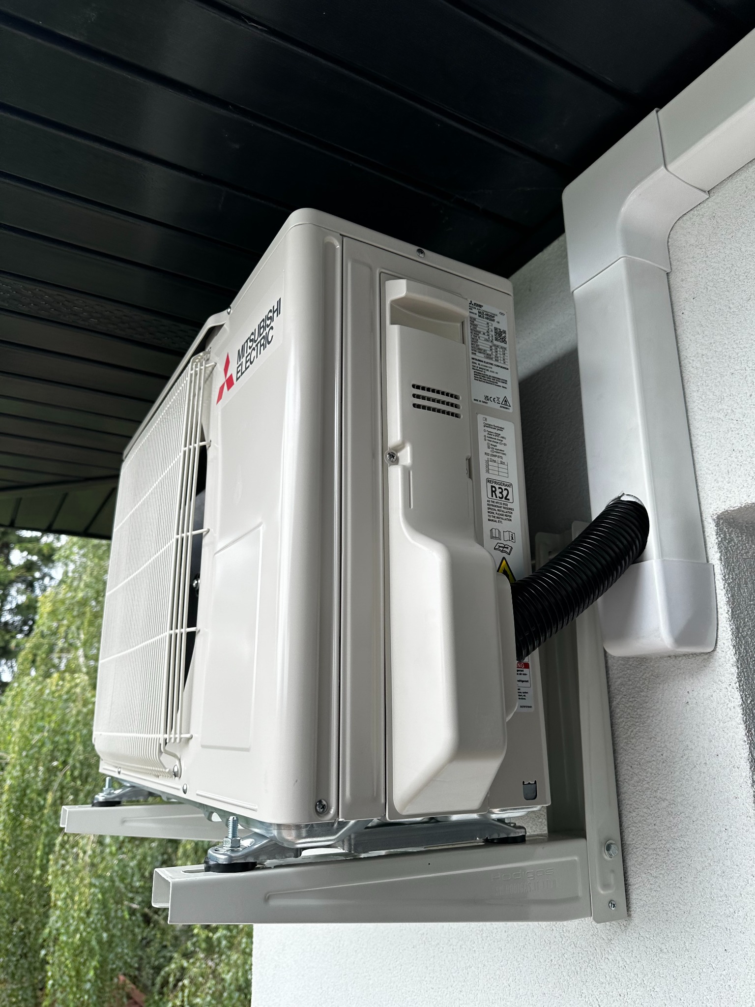 Zewnętrzna jednostka klimatyzacji Mitsubishi Electric zamontowana na wspornikach na elewacji budynku, z widocznym odprowadzeniem skroplin i fragmentem czarnego podbitki dachowej.