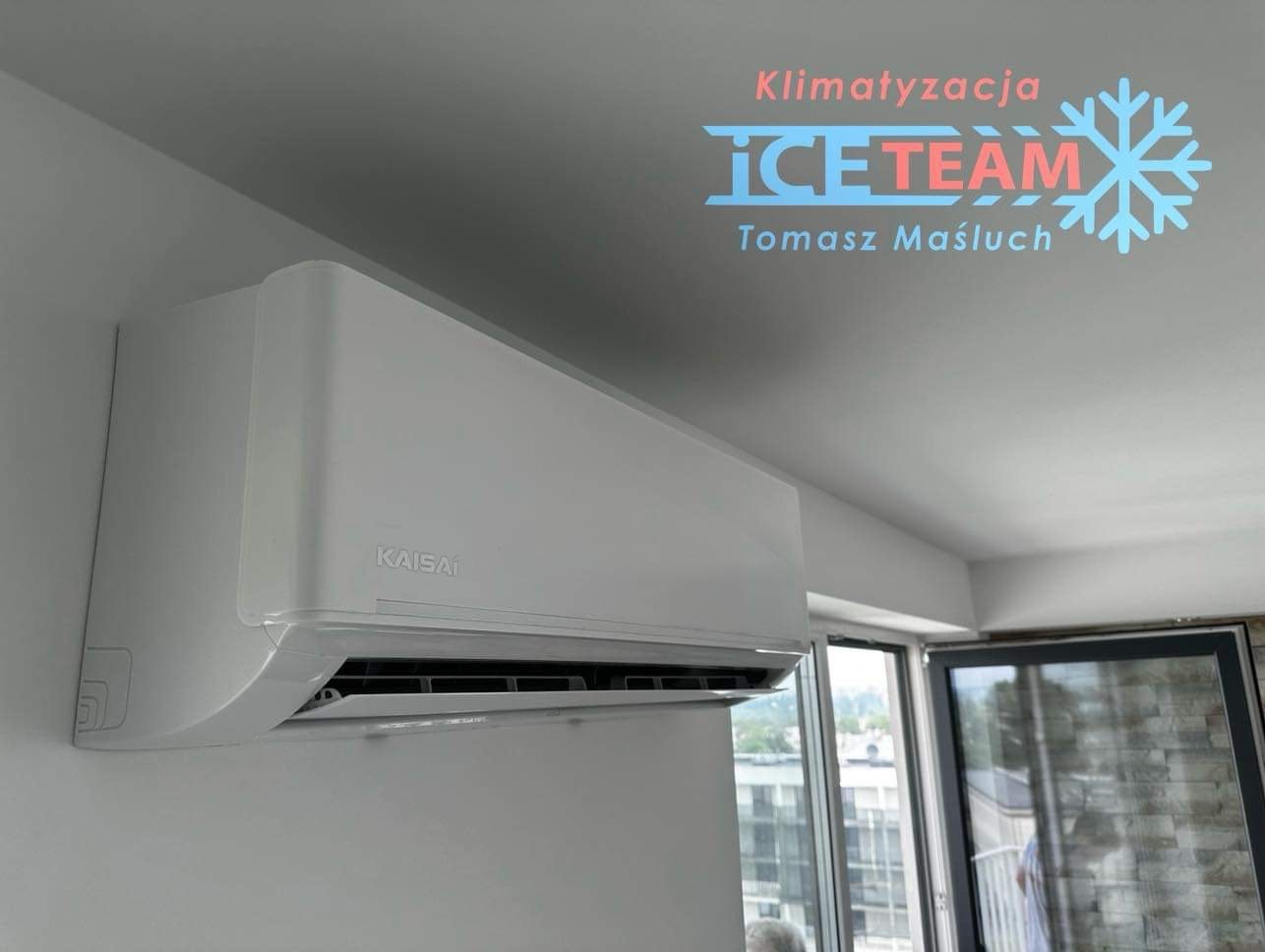 Biała klimatyzacja Kaisai zamontowana na ścianie w jasnym pomieszczeniu, widoczne logo firmy 'Ice Team' na suficie.