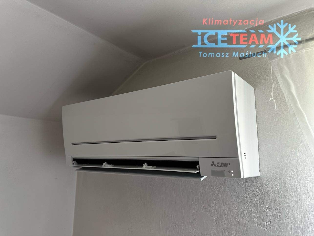 Biała jednostka klimatyzacji Mitsubishi Electric zamontowana na szarej ścianie w pomieszczeniu. Widoczny fragment sufitu i okna z firanką. W prawym górnym rogu logo firmy Klimatyzacja ICE Team.