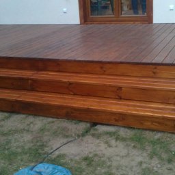 DREW-BUD Stolarstwo - Nowo wybudowany taras z drewna z trzema stopniami, widok z niskiej perspektywy na tle białej ściany budynku z oknem.