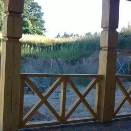 DREW-BUD Stolarstwo - Drewniana balustrada tarasu z krzyżowymi elementami, surowe drewno, widok na skarpę z zielenią i siatką ogrodzeniową w tle.