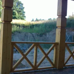 Drewniana balustrada tarasu z krzyżowymi elementami, surowe drewno, widok na skarpę z zielenią i siatką ogrodzeniową w tle.