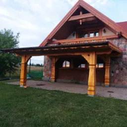 Drewniana konstrukcja zadaszenia tarasu przy domu z kamienną elewacją i czerwoną dachówką, widok na ogród i niebo.