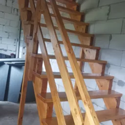 Jasne, drewniane schody z poręczą, wykonane z drewna o widocznym usłojeniu, na tle ściany z białych bloczków.