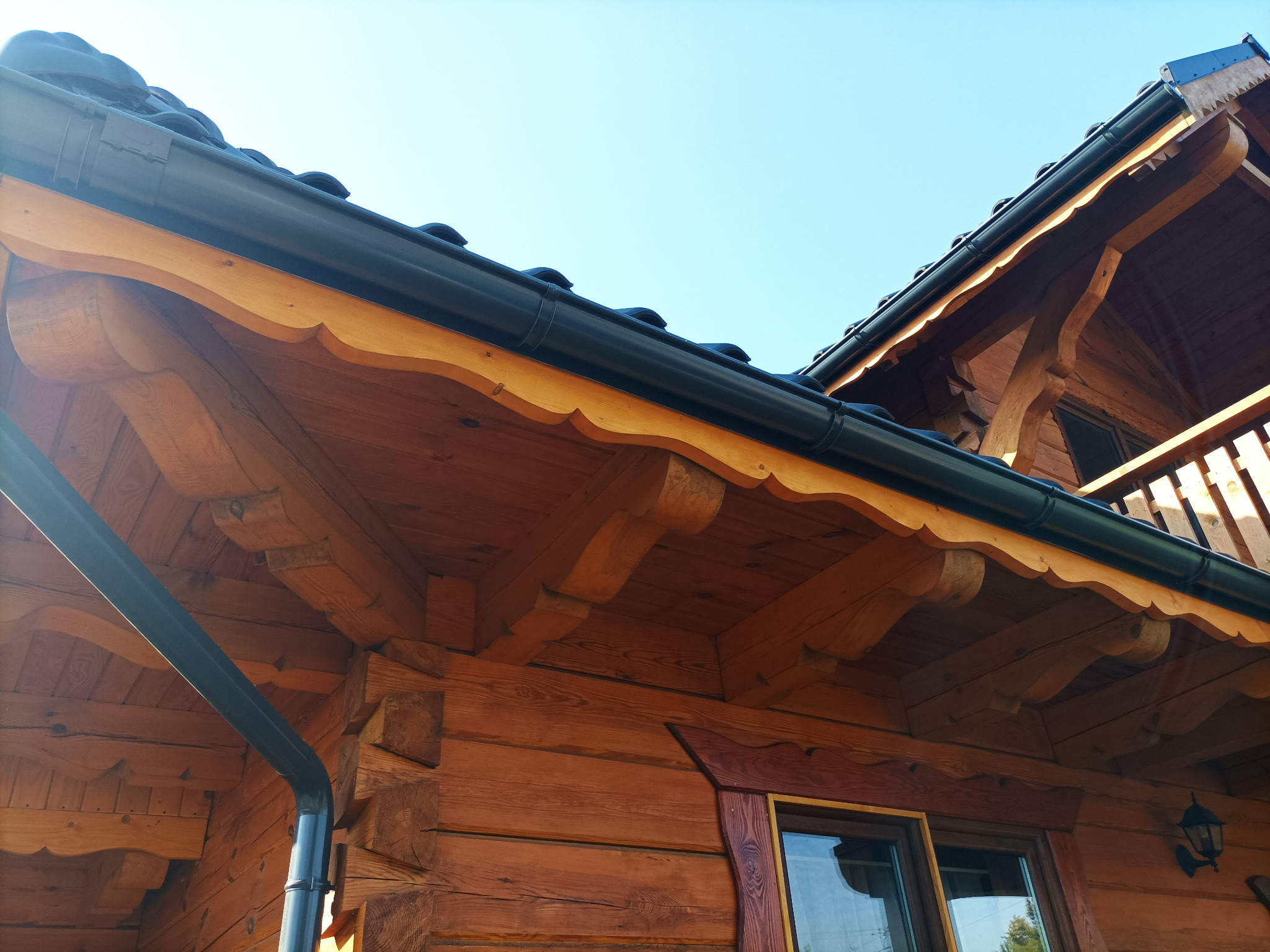 Fragment drewnianego domu z widocznym orynnowaniem, ozdobnym podbitkiem i fragmentem balkonu na tle błękitnego nieba.