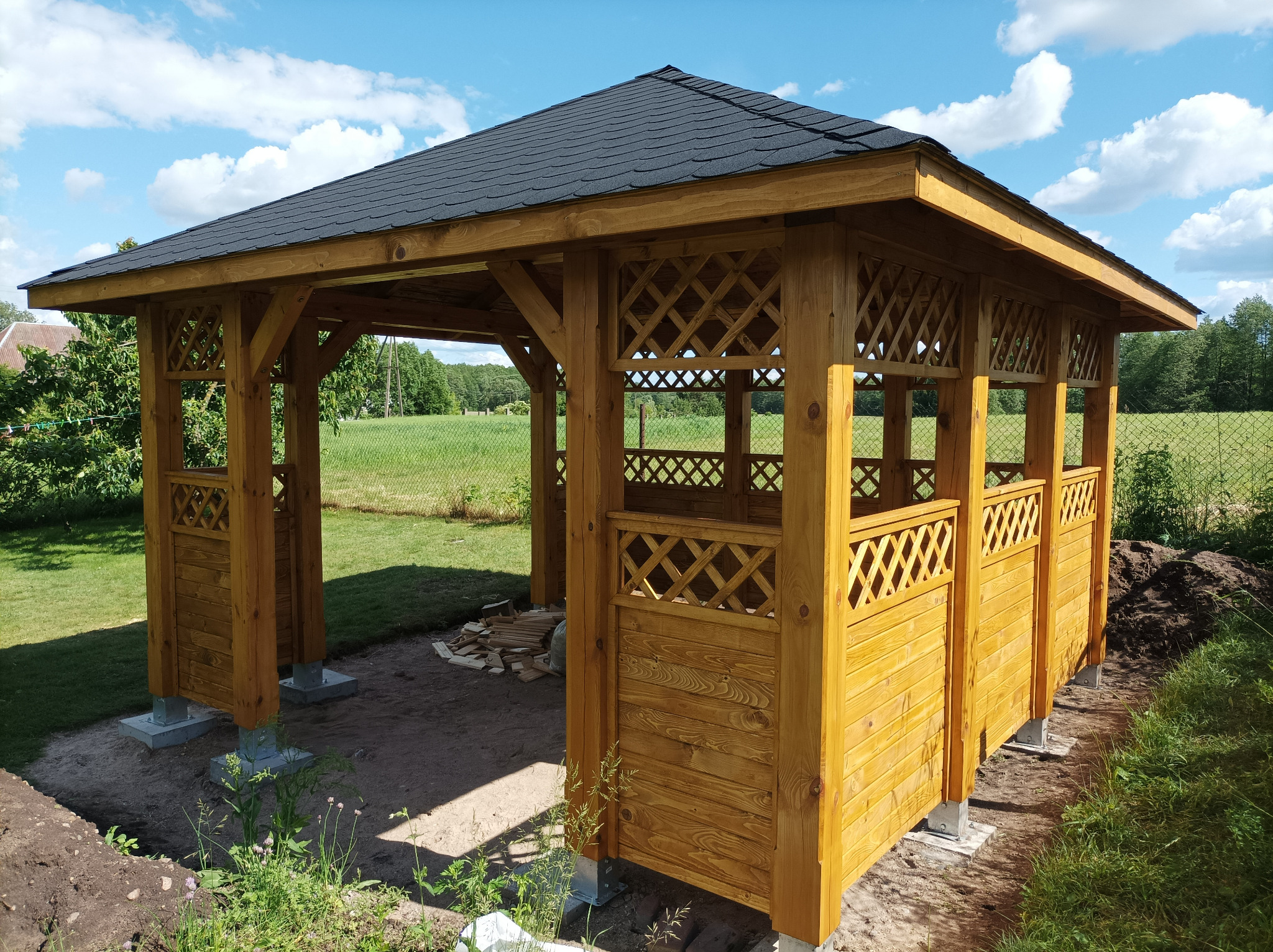 Drewniana altana z czarnym dachem w trakcie budowy, widoczne betonowe podstawy, w tle zielone pole i drzewa pod błękitnym niebem.