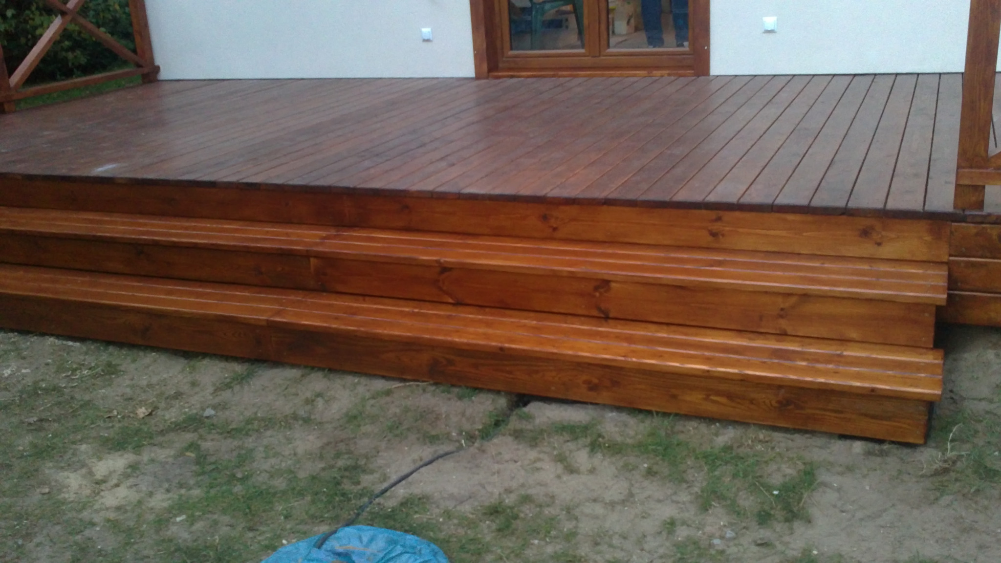 Nowo wybudowany taras z drewna z trzema stopniami, widok z niskiej perspektywy na tle białej ściany budynku z oknem.