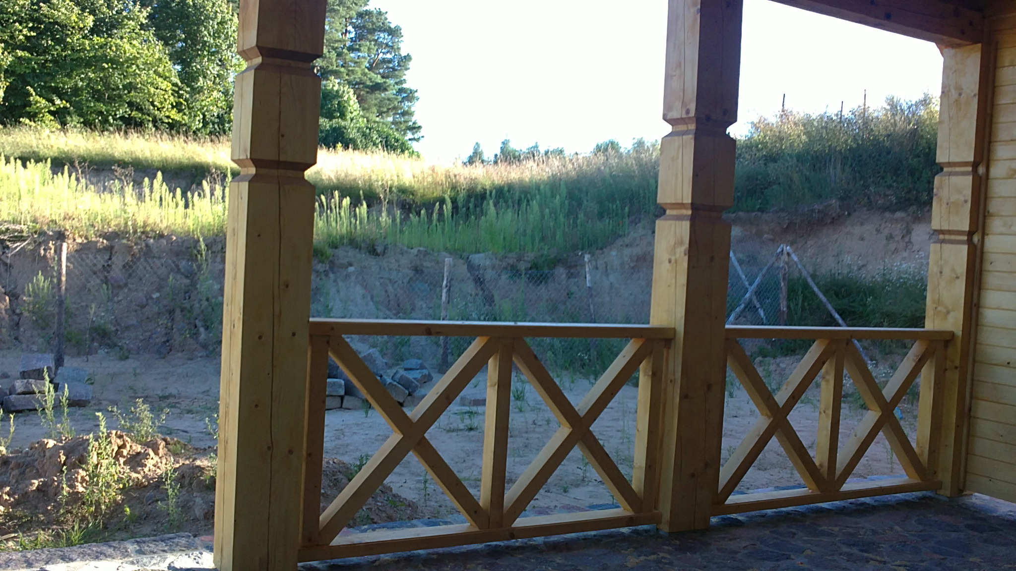 Drewniana balustrada tarasu z krzyżowymi elementami, surowe drewno, widok na skarpę z zielenią i siatką ogrodzeniową w tle.