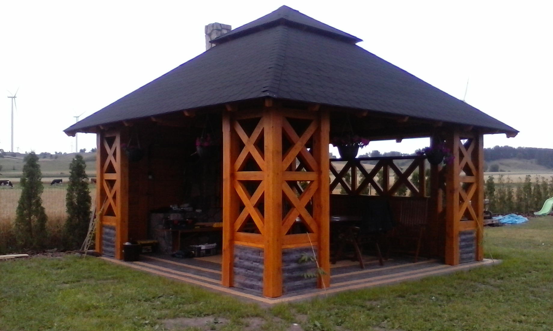 Drewniana pergola z ciemnym dachem, ozdobnymi drewnianymi słupami w kształcie krzyży i doniczkami z kwiatami, widok na pole z turbinami wiatrowymi w tle.