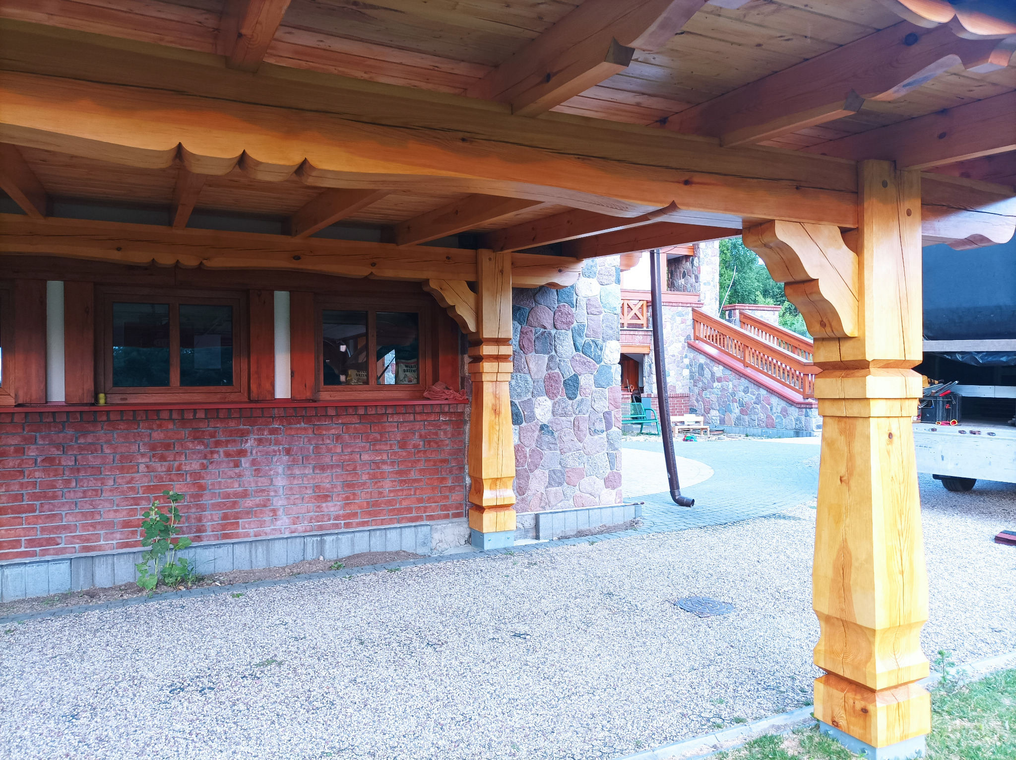 Drewniana konstrukcja z ozdobnymi wykończeniami, wsparta na kolumnach, z widocznym fragmentem kamiennej ściany i schodów w tle, oświetlona naturalnym światłem.