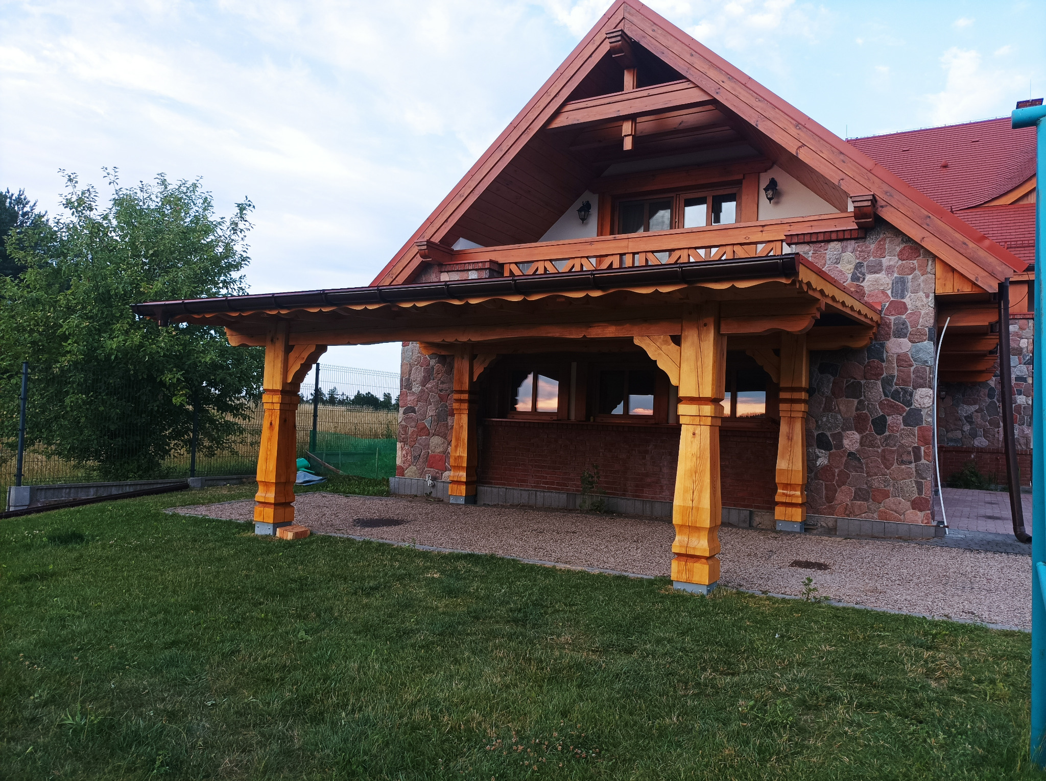 Drewniana konstrukcja zadaszenia tarasu przy domu z kamienną elewacją i czerwoną dachówką, widok na ogród i niebo.