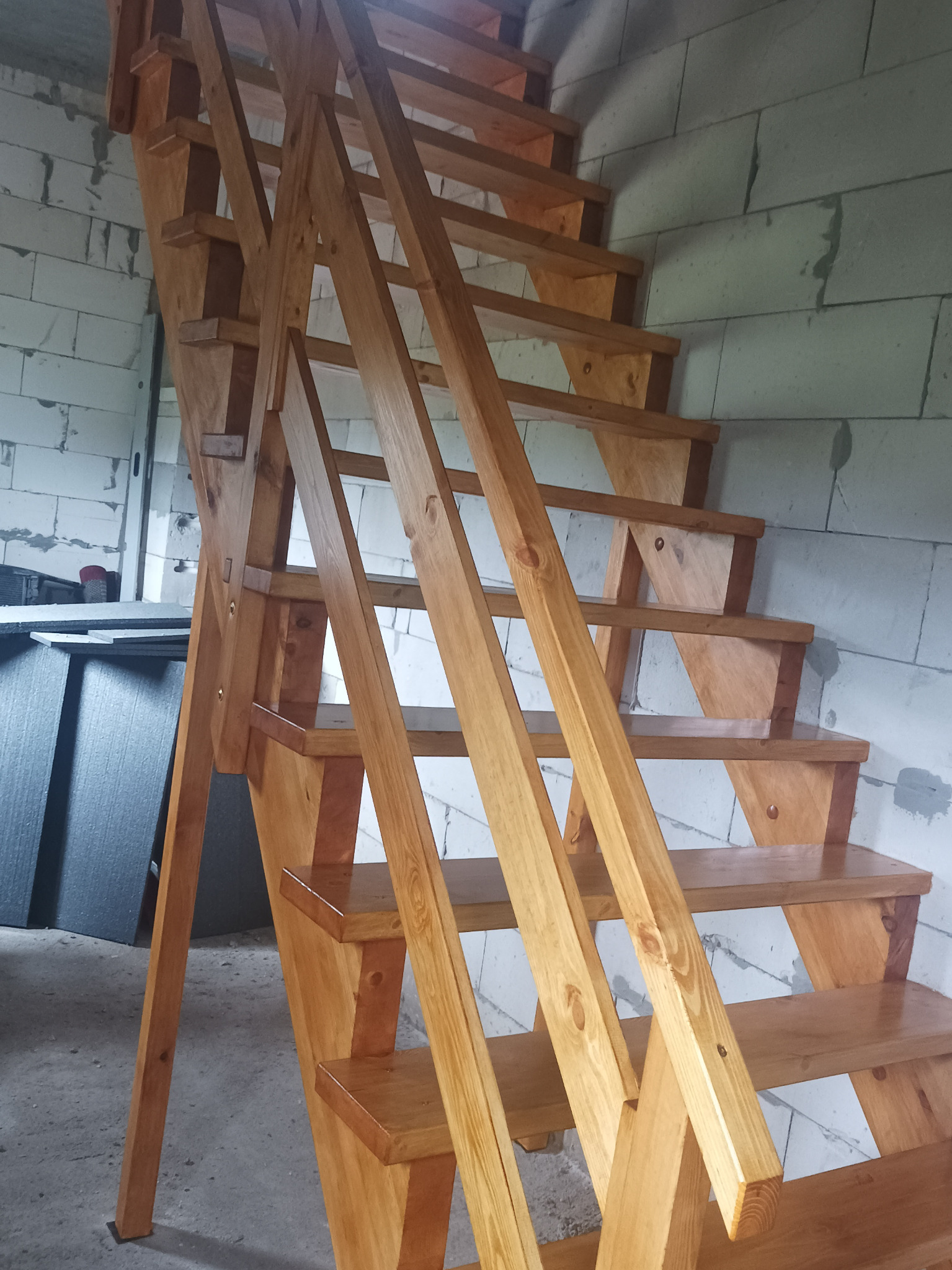 Jasne, drewniane schody z poręczą, wykonane z drewna o widocznym usłojeniu, na tle ściany z białych bloczków.