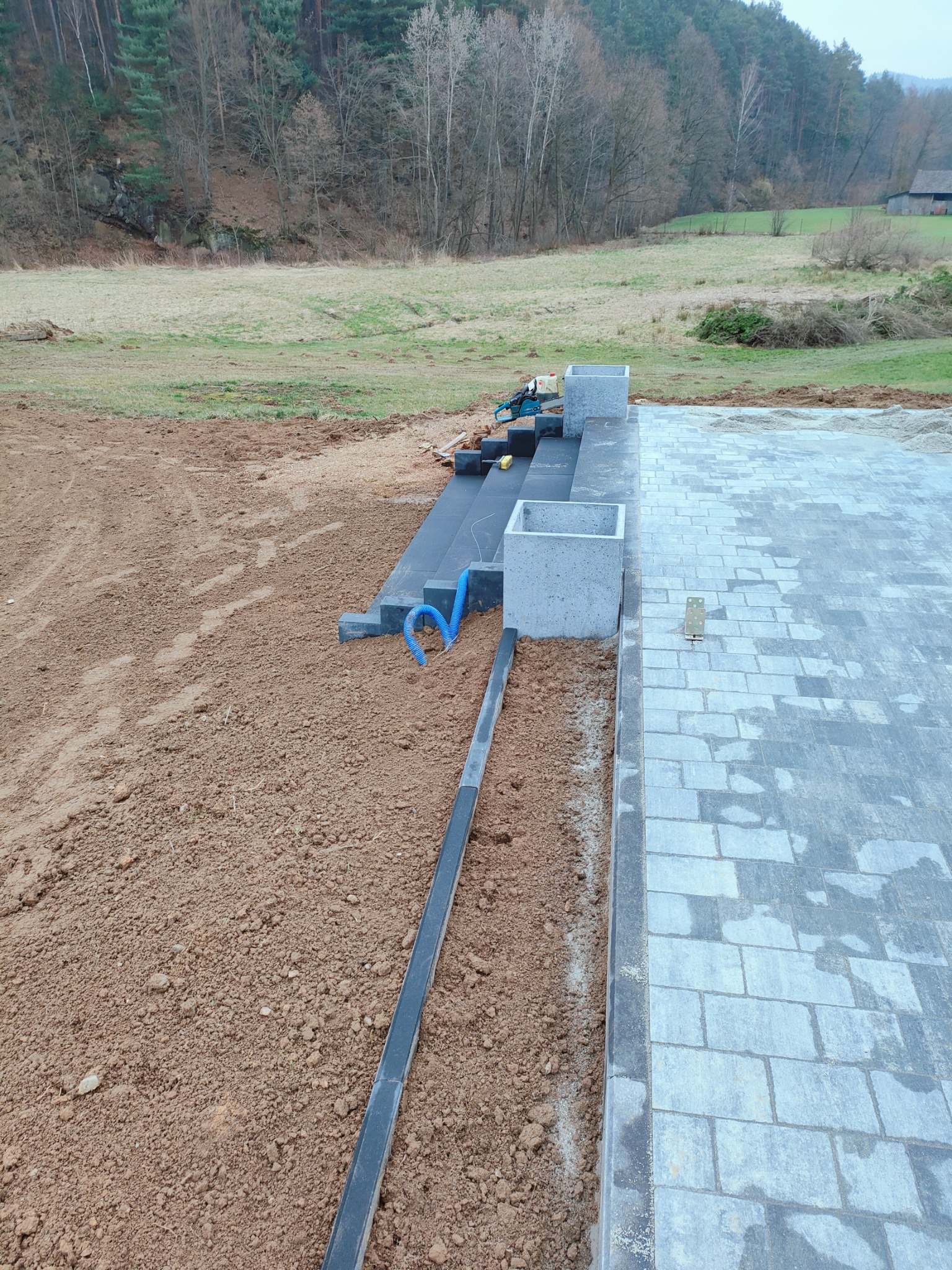 Układanie szarej kostki brukowej na zewnątrz z widocznymi elementami drenażu, niezakończony etap prac, otoczenie: ziemia i drzewa.