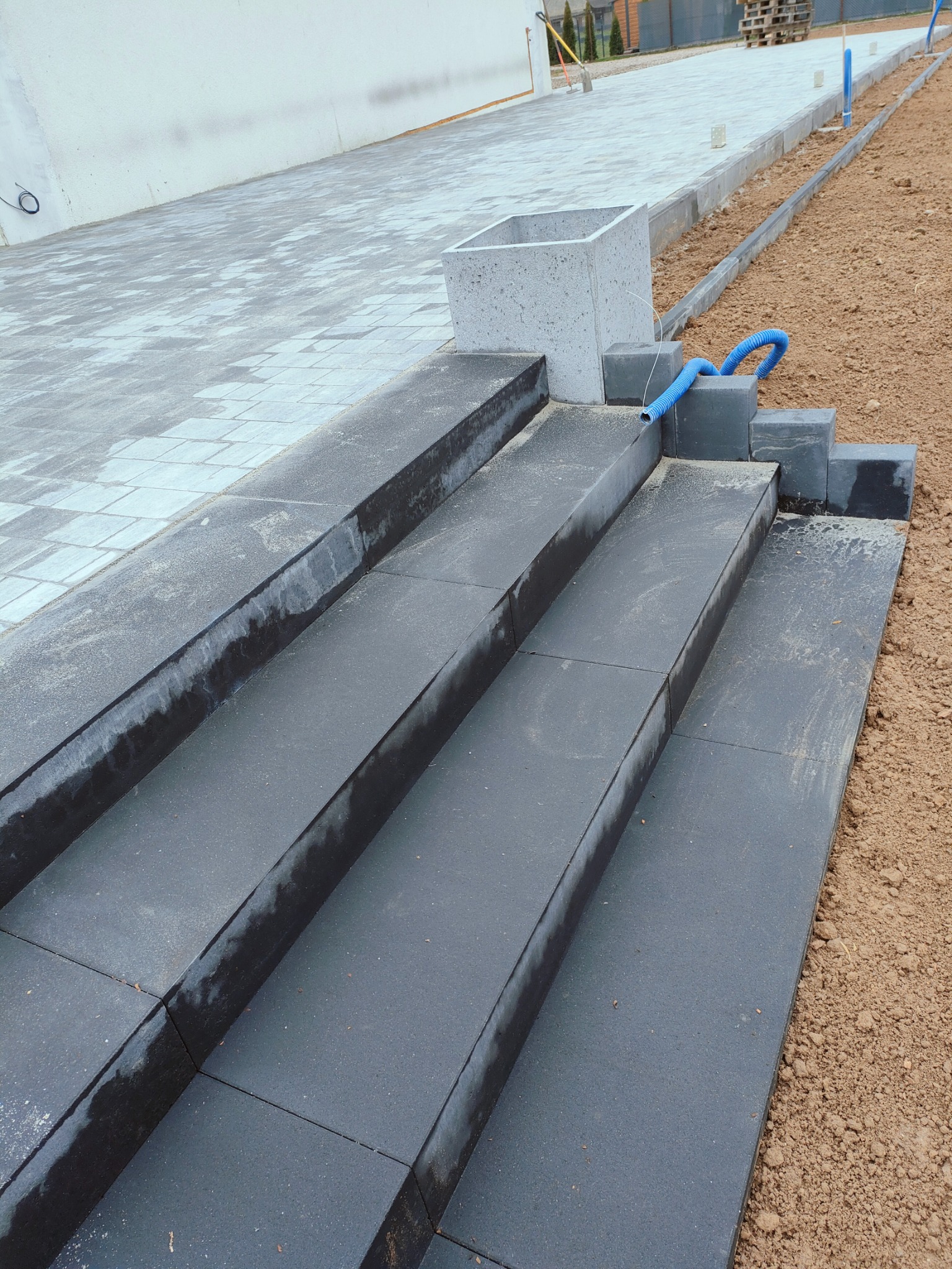 Nowe, ciemnoszare schody z kostki brukowej prowadzące do jasnoszarego chodnika, obok świeżo usypana ziemia i niebieski przewód.