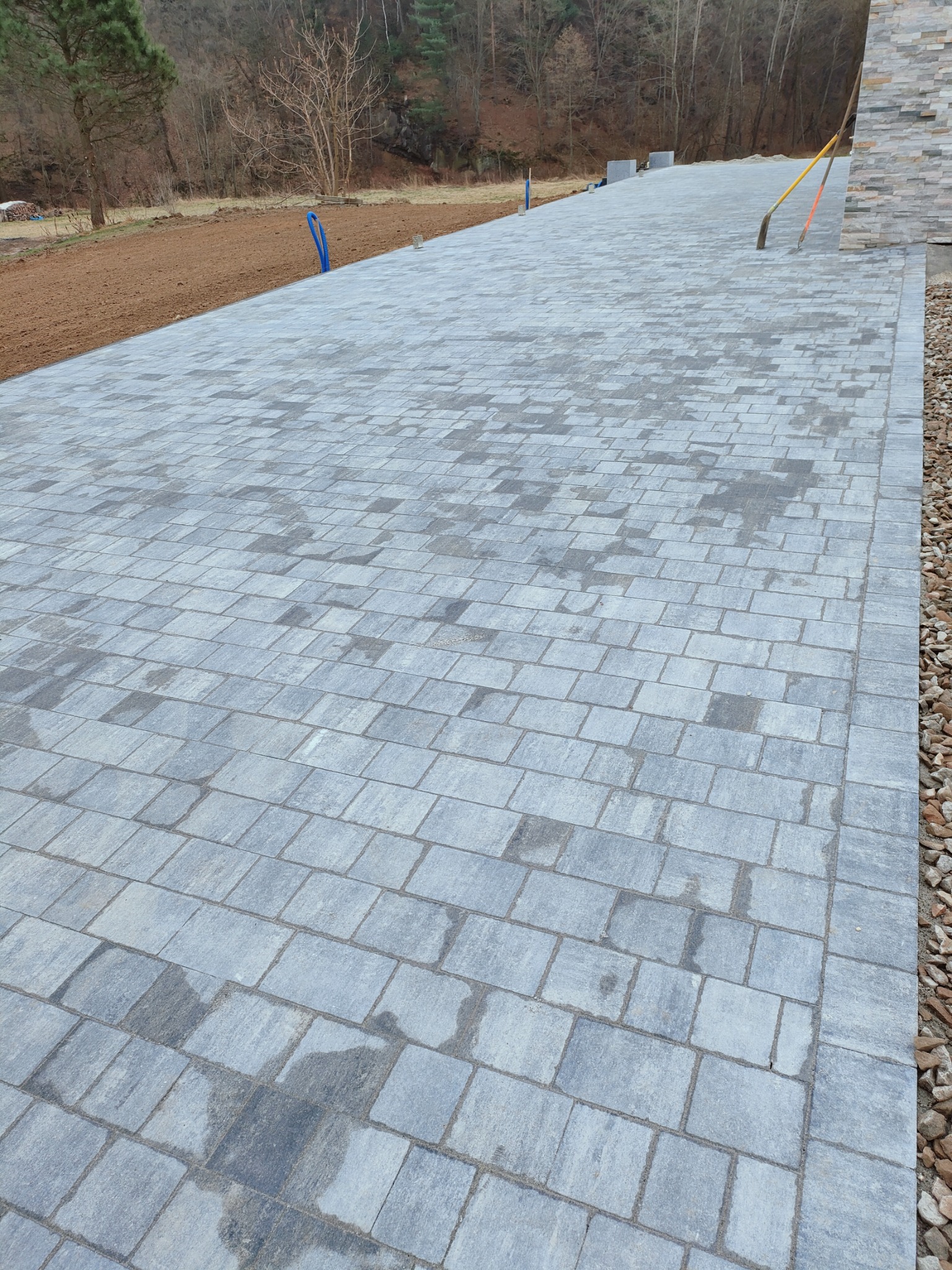 Ułożona szara kostka brukowa tworząca długą ścieżkę, obok widoczny pas żwiru i przygotowany teren pod trawnik, w tle drzewa i fragment budynku z kamienną elewacją. Dwie łopaty oparte o ścianę.