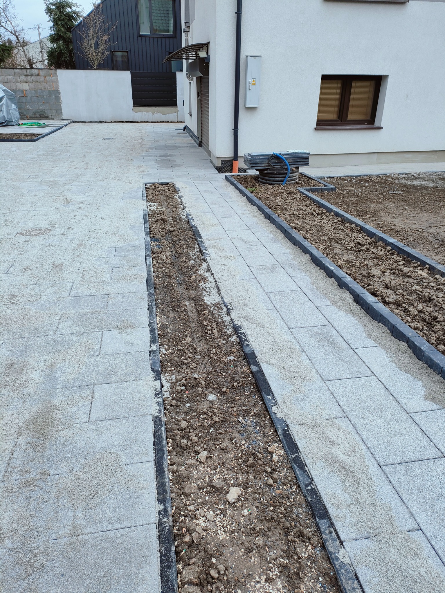 Ułożona ścieżka z jasnoszarej kostki brukowej wzdłuż białego domu, z przygotowanymi korytkami wypełnionymi ziemią po bokach, czekającymi na dalsze prace wykończeniowe.