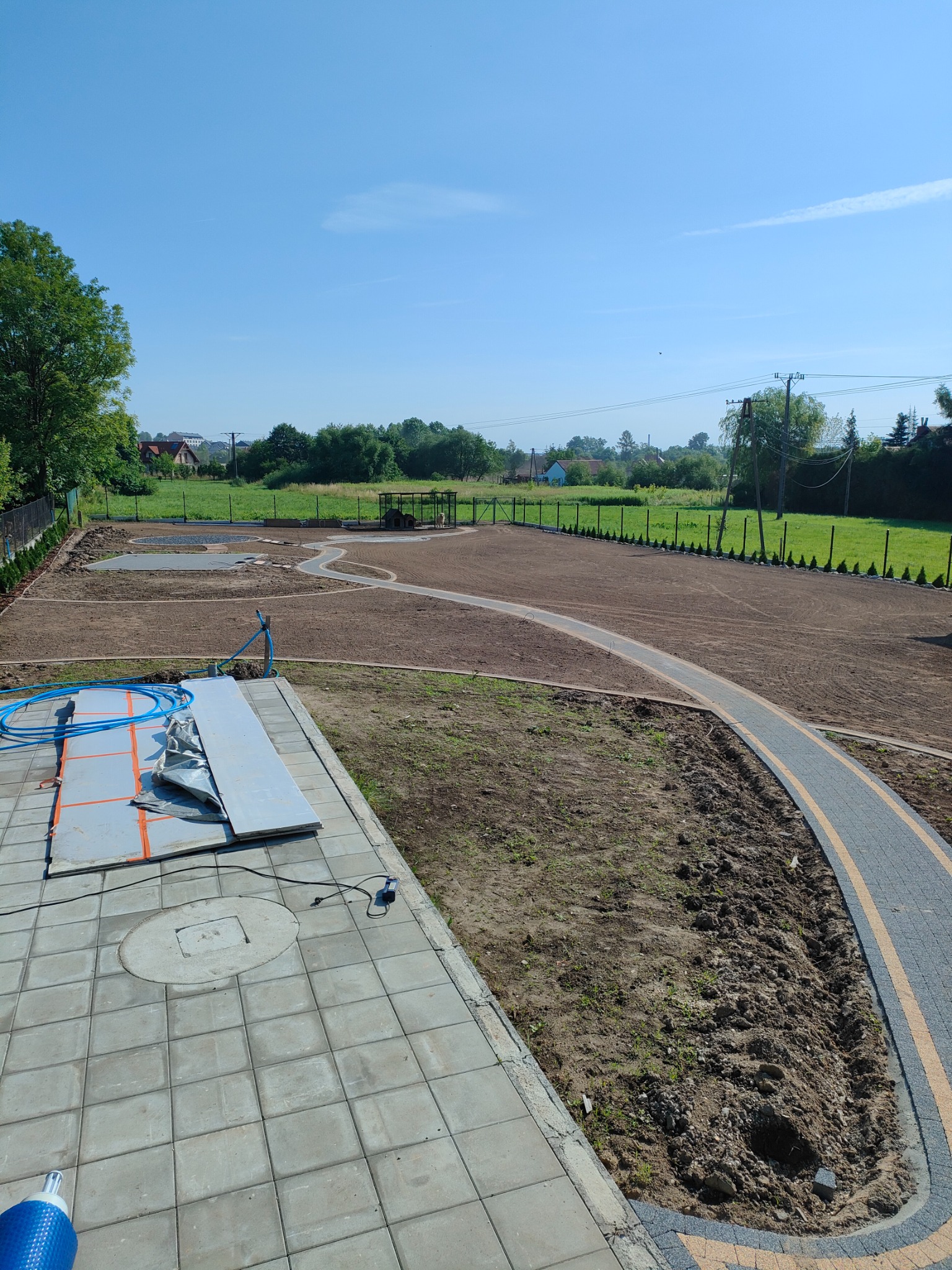 Przygotowany teren pod budowę z wytyczonymi ścieżkami z kostki brukowej, widoczne fundamenty i materiały budowlane oraz błękitne węże na kostce, tło stanowią drzewa i zabudowania.