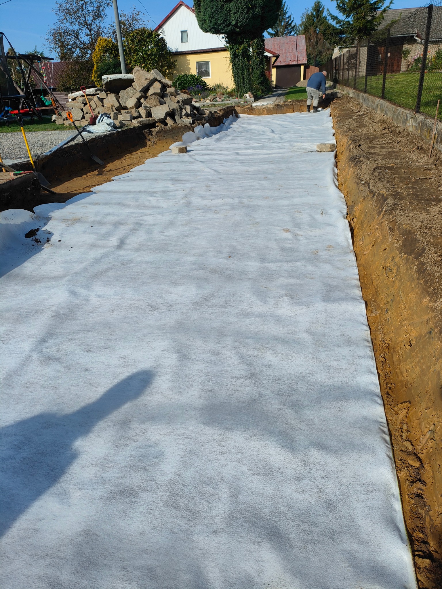 Przygotowany wykop pod budowę, wyłożony białą geowłókniną, widoczny fragment osoby pracującej oraz sterta kamieni w tle.