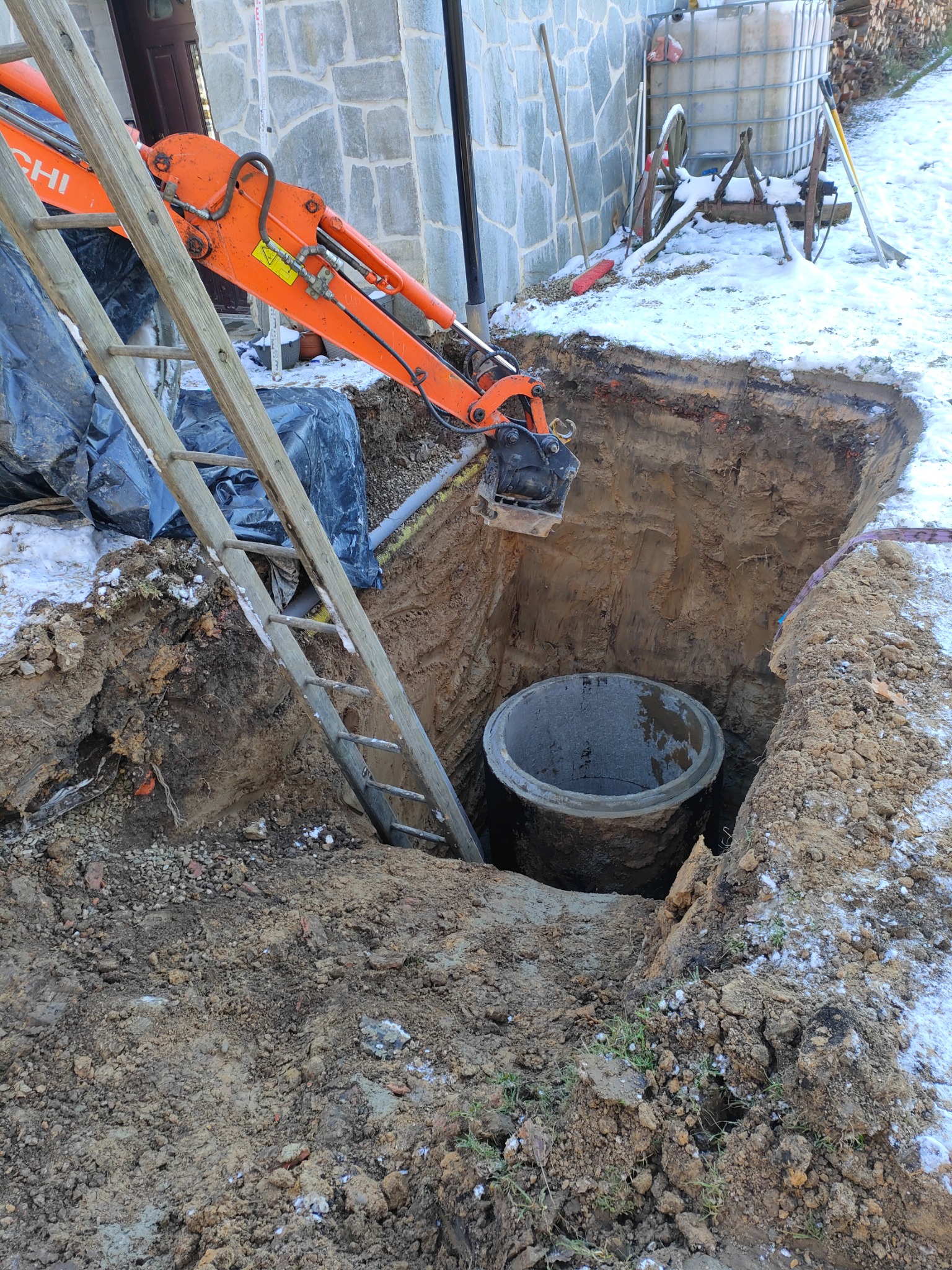 Pomarańczowa koparka Hitachi nad wykopem z betonowym kręgiem, drabina oparta o skarpę, resztki śniegu na ziemi, w tle budynek z kamienia.