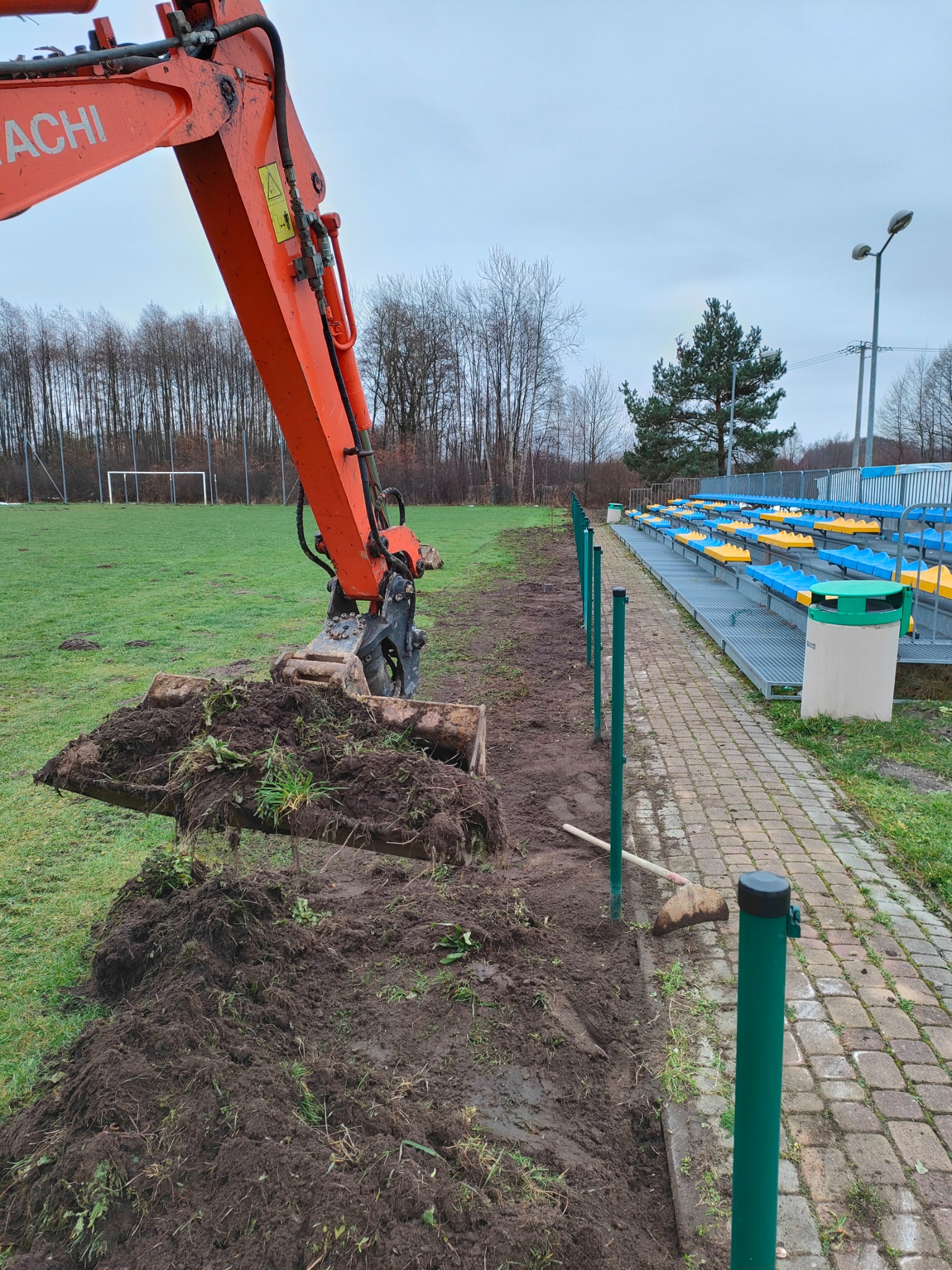Pomarańczowa koparka Hitachi usuwa warstwę ziemi z trawy obok trybun stadionu, przygotowując teren pod przyszłe prace.