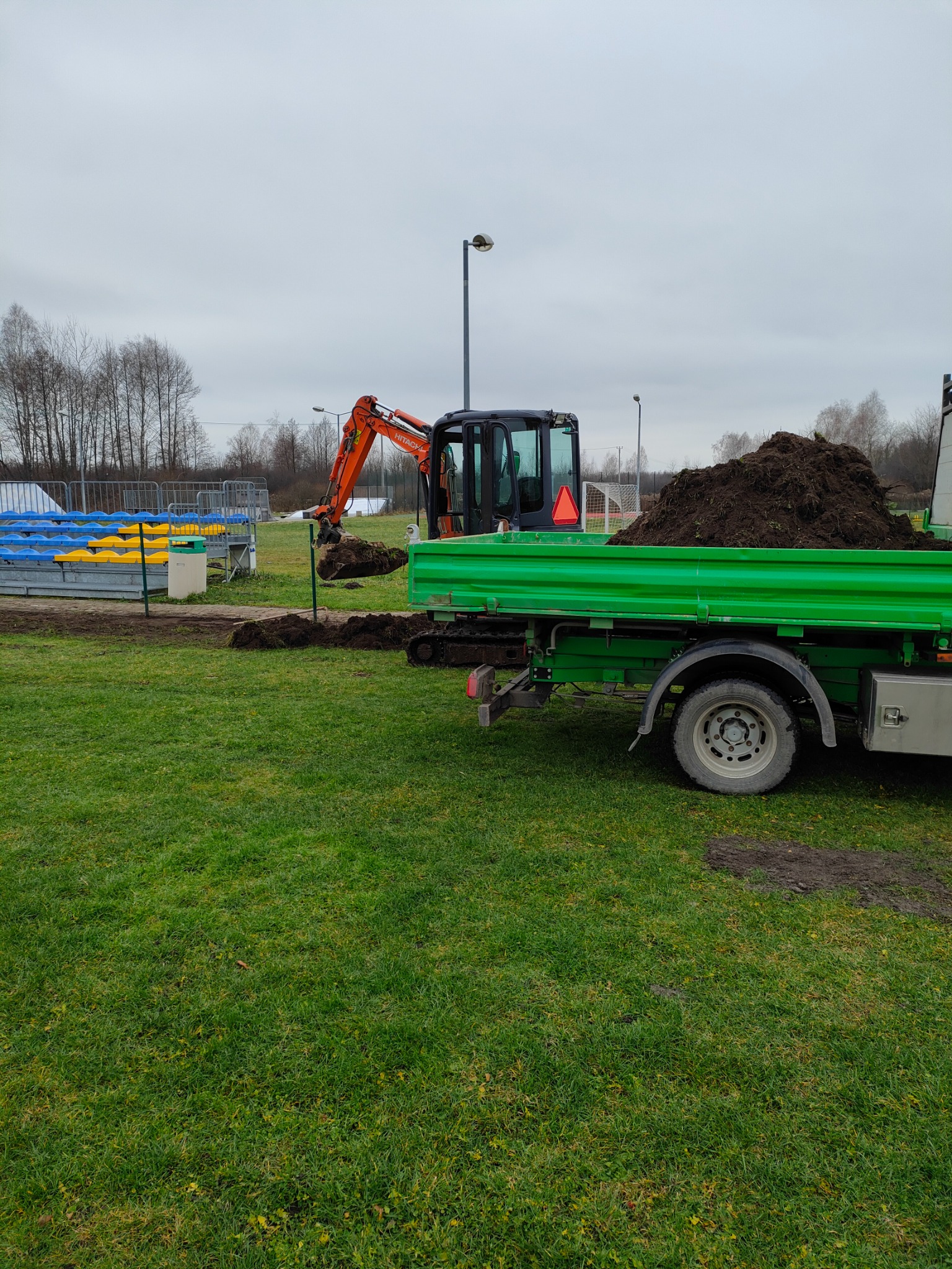 Mała koparka Hitachi nabiera ziemię i wrzuca ją na skrzynię zielonej ciężarówki na tle trawiastego terenu i trybun sportowych