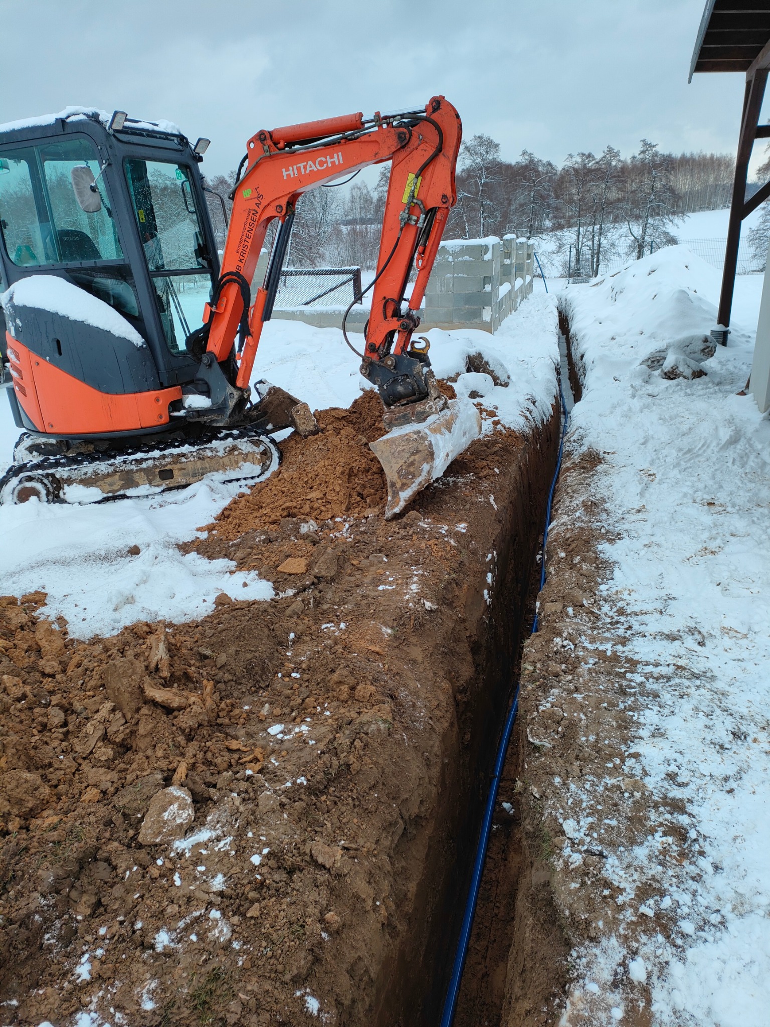 Pomarańczowa mini koparka Hitachi przy wykopie z ułożoną niebieską rurą w zasypanym śniegiem terenie.