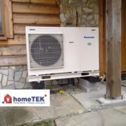 Zewnętrzna jednostka pompy ciepła Panasonic, zamontowana na betonowym fundamencie przy drewnianym domu z kamienną podmur&oacute;wką, widoczny fragment elewacji i drewniany słup werandy, logo firmy HomeTEK...
