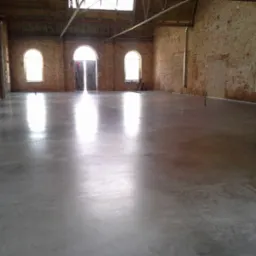 Gładka, szara powierzchnia w hali z ceglanymi ścianami i łukowymi oknami, refleksy światła podkreślają równość podłoża.