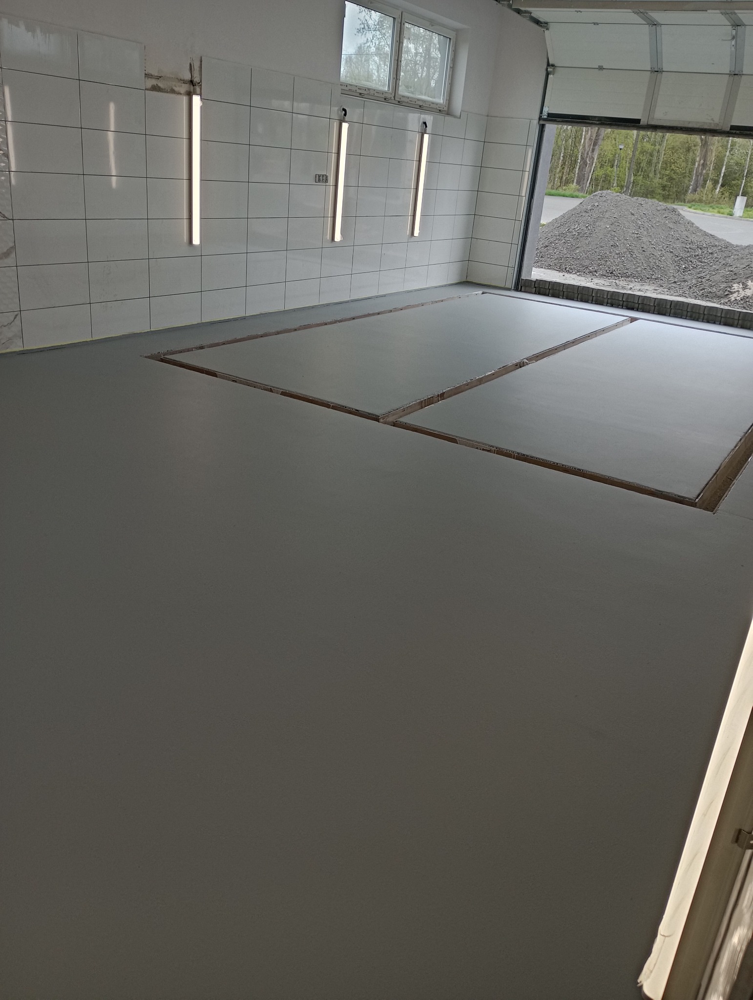 Świeżo wylana, szara powierzchnia w hali z widocznymi przerwami dylatacyjnymi, ściany wyłożone białymi płytkami, oświetlenie jarzeniowe, otwarte wrota garażowe z widokiem na hałdę żwiru.