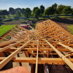 Drewniana konstrukcja dachu w trakcie budowy, widok z góry na tle zielonej łąki i drzew, częściowo pokryta cegłami.