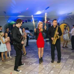 Występ na żywo: kobieta w czerwonej sukience trzyma zimne ognie, obok mężczyźni z saksofonem i gitarą elektryczną, w tle tańczący goście na sali udekorowanej płatkami róż.