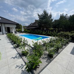 KB Konrad Borzym - Nowoczesny taras z basenem, wyłożony jasnoszarą kostką. Geometryczny układ, rośliny wzdłuż krawędzi, dom w tle. Słoneczny dzień, błękitna woda.