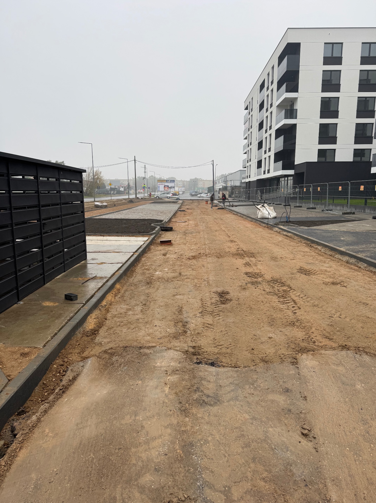 Przygotowanie podłoża pod kostkę brukową na terenie inwestycji deweloperskiej. Widoczne obrzeża, wyrównany teren i częściowo ułożona warstwa podsypki. Szara, pochmurna pogoda.