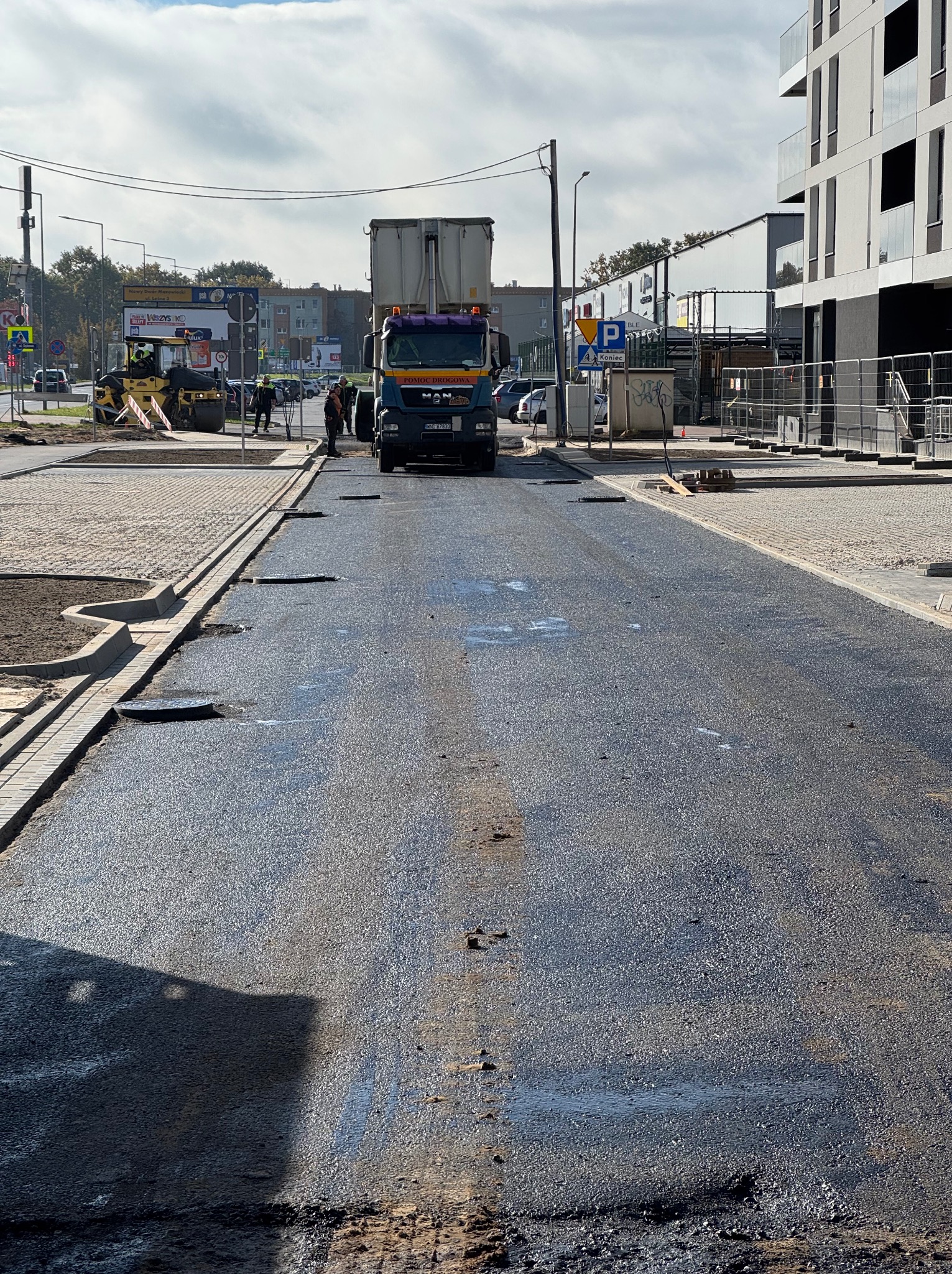 Świeżo wylany asfalt na drodze z widocznym walcem i ciężarówką MAN w tle, obrzeża z kostki brukowej. Prace drogowe w trakcie realizacji.