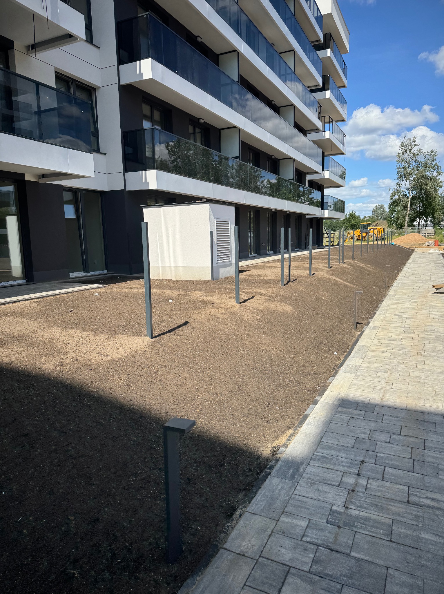 Przygotowany teren pod trawnik z zainstalowanym oświetleniem wzdłuż ścieżki z kostki brukowej, na tle nowoczesnego budynku apartamentowego.