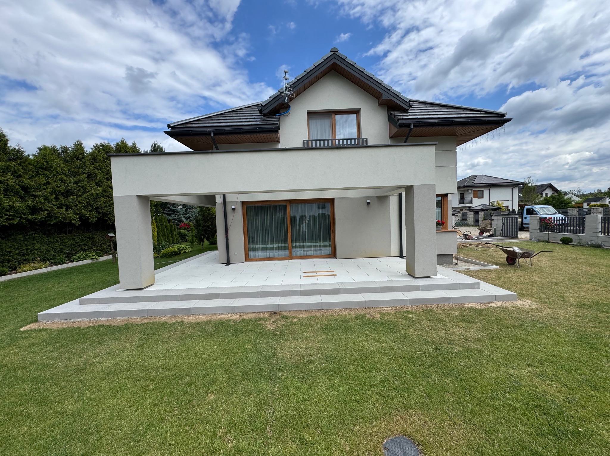 Nowoczesny taras z jasnoszarej kostki przy domu z dużymi oknami i zielonym trawnikiem. Widoczny wózek budowlany w tle. Słoneczny dzień.