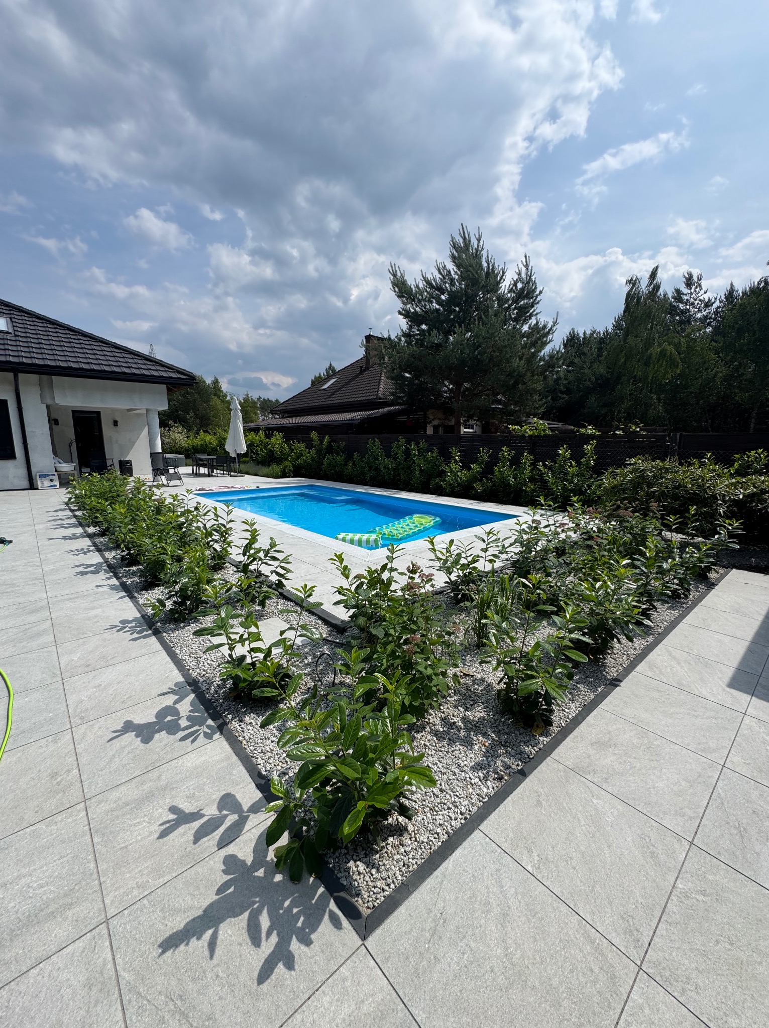 Nowoczesny taras z basenem, wyłożony jasnoszarą kostką. Geometryczny układ, rośliny wzdłuż krawędzi, dom w tle. Słoneczny dzień, błękitna woda.