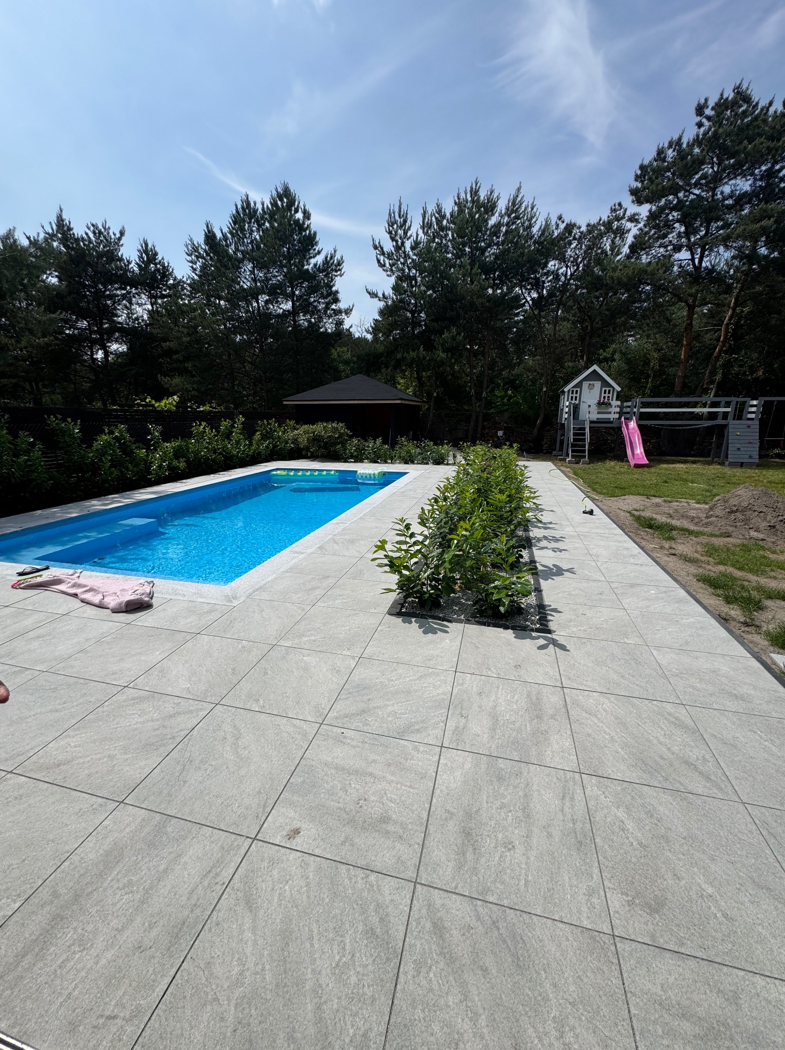 Szara, duża kostka brukowa wokół basenu, z rzędem zielonych krzewów i widokiem na domek dla dzieci oraz altanę w tle, w słoneczny dzień.