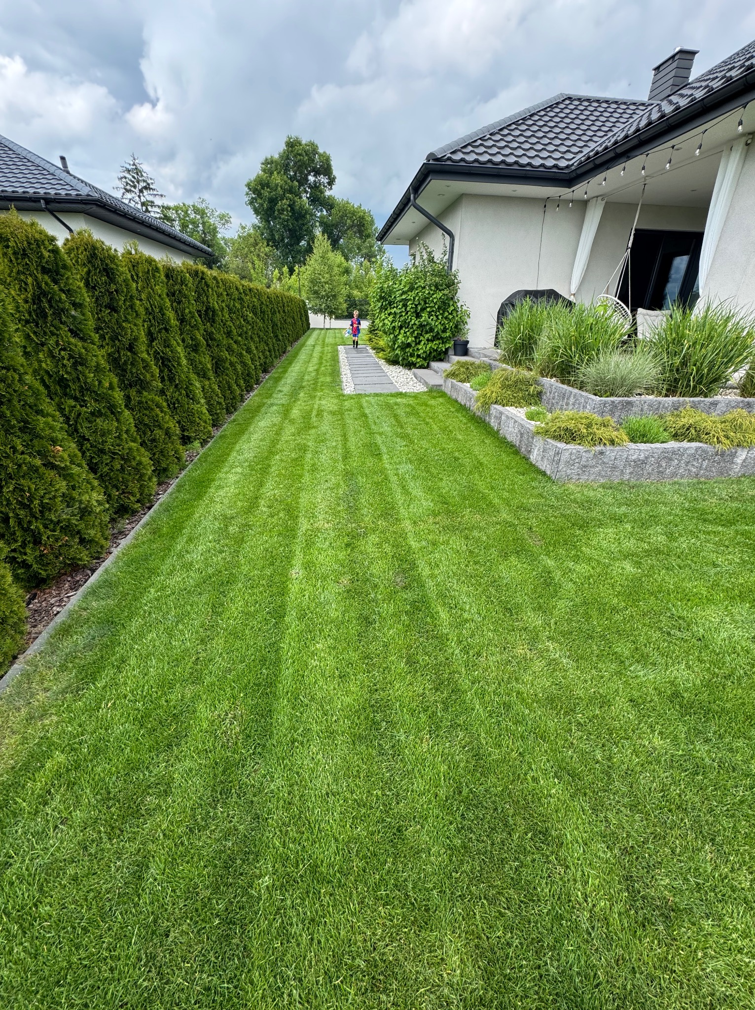 Zadbany trawnik z równo przystrzyżoną trawą, obrzeżami z kostki i żywopłotem z tui. W tle dom z elementami dekoracyjnymi i osoba na ścieżce z płyt.