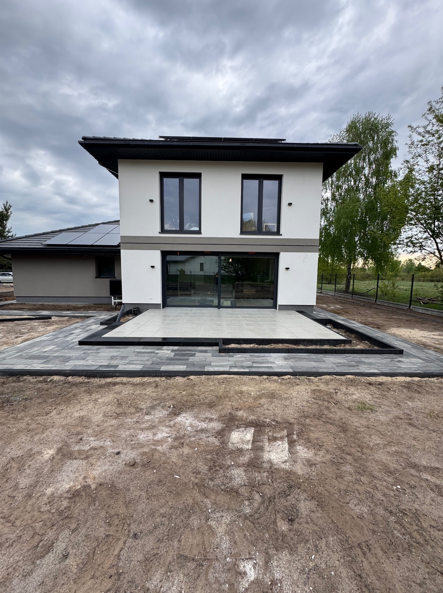 Nowoczesny dom z tarasem wyłożonym jasnymi płytami i obrzeżem z ciemnej kostki. Widoczne ułożone ścieżki z kostki brukowej. Pochmurne niebo w tle.