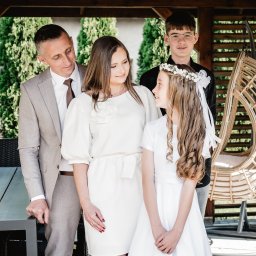 Wioleta Grzywińska - Rodzinne zdjęcie w altanie: ojciec w garniturze, matka w białej sukience, syn w okularach i córka w białej sukience komunijnej z wiankiem, wszyscy uśmiechnięci.