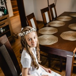 Wioleta Grzywińska - Uśmiechnięta dziewczynka w białej sukience komunijnej i wianku siedzi przy stole z plecionymi podkładkami, patrząc w górę.