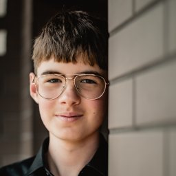 Wioleta Grzywińska - Portret młodego mężczyzny w okularach z metalowymi oprawkami, opierającego się o ceglaną ścianę, ubrany w ciemną koszulę.