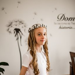 Wioleta Grzywińska - Portret dziewczynki w białej sukience i wianku z białych kwiatów na głowie, na tle ściany z motywem dmuchawca i napisem, delikatny uśmiech.