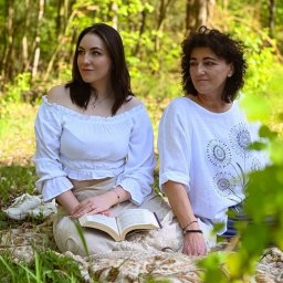 Wioleta Grzywińska - Dwie kobiety w białych bluzkach siedzą na kocu w lesie. Młodsza kobieta trzyma otwartą książkę na kolanach, obie spoglądają w lewo.