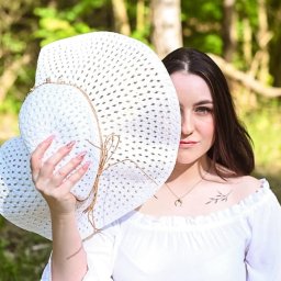 Wioleta Grzywińska - Portret kobiety z białym kapeluszem zakrywającym część twarzy, w tle rozmyta zieleń drzew.