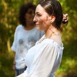 Wioleta Grzywińska - Portret profilowy młodej kobiety w białej bluzce z odkrytymi ramionami, z włosami upiętymi w kok z ozdobną gumką, na tle zamazanego, zielonego tła z drugą osobą w tle.