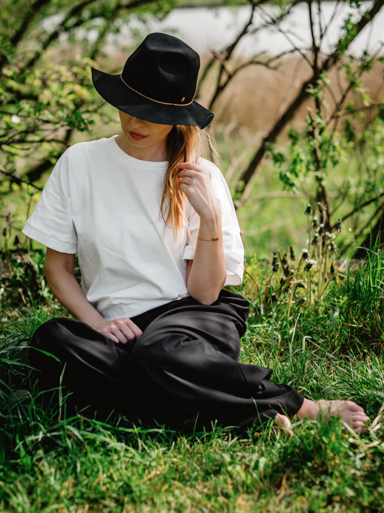 Kobieta w czarnym kapeluszu siedzi boso na trawie, w tle zieleń drzew i krzewów, naturalne światło.